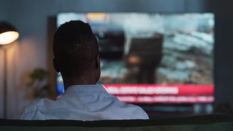 Back View of Black Man Sitting on a Sofa and Watching TV with Live News During the Evening. Entertainment and Technologies Concept. Back View Moving Shot with Out of Focus Blurred TV Screen