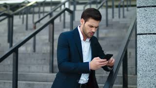 Close up serious businessman standing with phone in hand at street. Disappointed man typing on phone outdoors at stairs